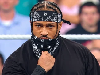 Nahaufnahme von Carmelo Hayes mit geflochtenen Haaren und schwarzem Bandana, der mit ernstem Blick in die Kamera schaut und seine Gesichtsmaske festhält. Im Hintergrund sind ein Ringseil und Zuschauer zu sehen.