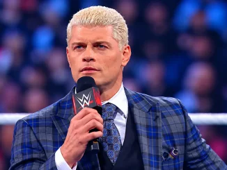 Cody Rhodes steht im WWE-Ring und hält ein Mikrofon in der Hand. Er trägt einen blau karierten Anzug mit passender Krawatte und blickt ernst in die Arena.
