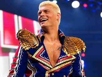 Cody Rhodes lächelt in einer blau-rot-weißen Jacke mit goldenen Schulterverzierungen vor einer beleuchteten WWE-Kulisse.