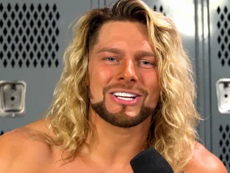 Lexis King mit langen blonden Haaren und Bart lächelt während eines Interviews vor grauen Umkleideschränken.