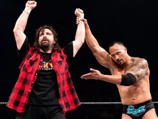 Mick Foley jubelt mit erhobenen Armen im WWE-Ring, während The Rock seinen Arm hochhält und auf ihn zeigt.