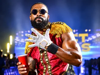 Montez Ford mit Sonnenbrille, rot-goldenem Ringoutfit und weißem Handschuh hält einen roten Becher während seines WWE-Einzugs.
