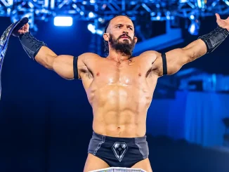 PAC hält mit ausgestreckten Armen einen Championship-Gürtel in seiner rechten Hand vor blau beleuchteter Arena-Kulisse.