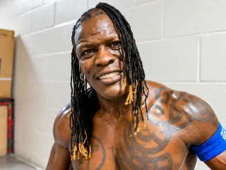 R-Truth mit langen Dreadlocks, tätowiertem Oberkörper und blauem Armband backstage.