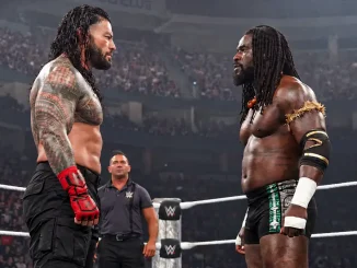 Roman Reigns und Oba Femi stehen sich im Wrestling-Ring gegenüber und blicken sich ernst an. Im Hintergrund steht ein WWE-Offizieller.