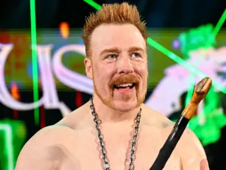 Sheamus lächelt mit einem Shillelagh und einer Kette um den Hals vor einem Hintergrund mit grünen Lichteffekten.