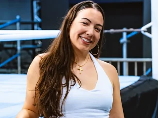 Sherilyn Guerrero lächelt in die Kamera vor einem Wrestling-Ring. Sie trägt ein hellblaues Oberteil, goldene Creolen und eine Halskette mit Kreuzanhänger.