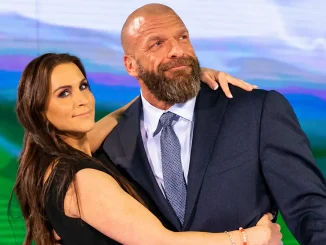 Stephanie McMahon umarmt Triple H, der im dunklen Anzug vor einem blau-grünen Hintergrund leicht nach oben blickt.