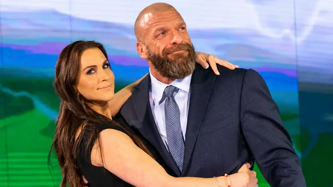 Stephanie McMahon umarmt Triple H, der im dunklen Anzug vor einem blau-grünen Hintergrund leicht nach oben blickt.