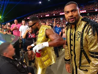 The Street Profits in goldfarbenen Outfits stehen bei WWE Raw neben einem sitzenden Sicherheitsmitarbeiter am Rand des Zuschauerbereichs.
