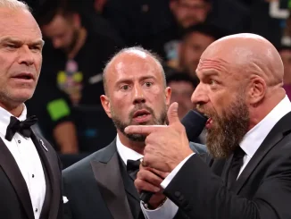 Triple H spricht mit Mikrofon und deutet auf Billy Gunn, während ein weiterer Mann im Anzug zwischen beiden steht und die Auseinandersetzung beobachtet.