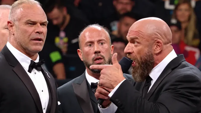 Triple H spricht mit Mikrofon und deutet auf Billy Gunn, während ein weiterer Mann im Anzug zwischen beiden steht und die Auseinandersetzung beobachtet.