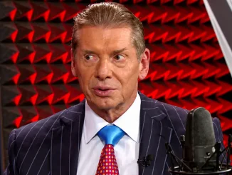 Vince McMahon im dunklen Nadelstreifenanzug mit blauer Krawatte vor einem Mikrofon und rot beleuchtetem Studiohintergrund.